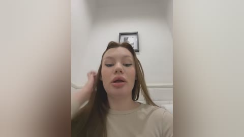 Media: Video of a young woman with long, straight brown hair, wearing a beige shirt, standing in a narrow, white-paneled hallway with a framed black-and-white picture on the wall.