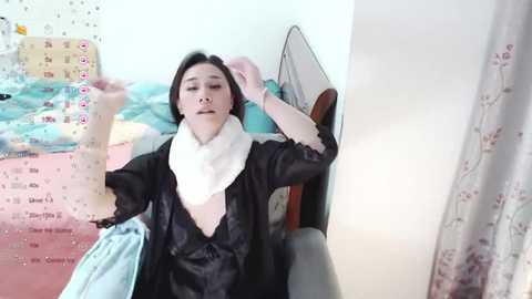 Media: A video of an Asian woman with short black hair, wearing a black satin robe and white scarf, sitting on a bed with a pink and white floral curtain, holding a blue bag, in a cozy, dimly lit room.