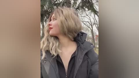 Video of a blonde woman with wavy hair, wearing a black puffer jacket, gazing upward in a park setting with tall trees and overcast sky.