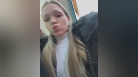 Media: Video of a young Caucasian woman with long blonde hair, wearing a white turtleneck and black jacket, puckering her lips in a playful manner. The background shows a blurred, dark interior.