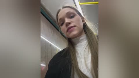 Media: Video of a young Caucasian woman with long blonde hair, wearing a black jacket over a white turtleneck, taken from a low angle in a dimly lit, industrial setting.