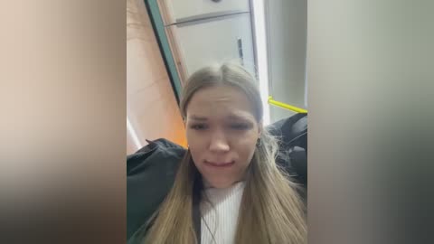 Media: Video of a young Caucasian woman with long blonde pigtails and light skin, wearing a white sweater, sitting on a train seat, surrounded by beige and green tones.