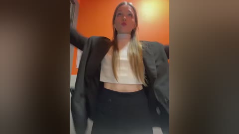 Media: Video of a blonde woman in a black blazer, white crop top, and black pants, leaning against a wall with orange and grey walls in the background.