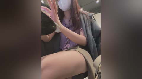 Media: Video of an East Asian woman with long brown hair, wearing a gray coat, purple blouse, and face mask, seated in a car, with blurry background and car interior details visible.