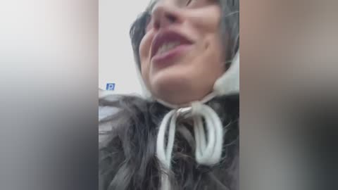 Media: A blurry video of a woman with a wide grin, wearing a gray hooded jacket with white fur trim, and a blue and white patterned scarf.