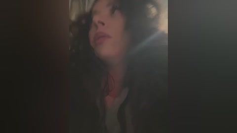 Media: A dimly lit video of a young woman with curly hair and light skin, wearing a black jacket and red scarf, gazing upward with a contemplative expression.