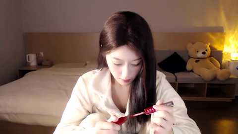 Media: A video shows a young Asian woman with long black hair, wearing a beige hoodie, flossing her teeth in a cozy bedroom with a white bed, a teddy bear, and a lit candle.