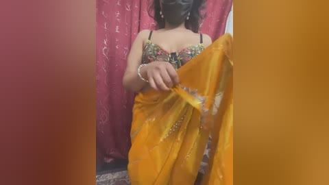 Media: Video of a South Asian woman in a yellow saree, black mask, and floral blouse, holding the saree with a silver bracelet. Background features a red curtain and yellow wall.