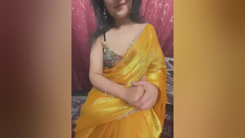 Media: A video of a South Asian woman in a yellow sari, with a floral blouse underneath, sitting against a red floral-patterned wall, wearing a bracelet.