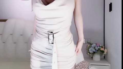 Media: A video of a woman in a white, sleeveless dress, adjusting her waistband, standing in a modern, minimalist bedroom with a bed, floral arrangement, and white walls.