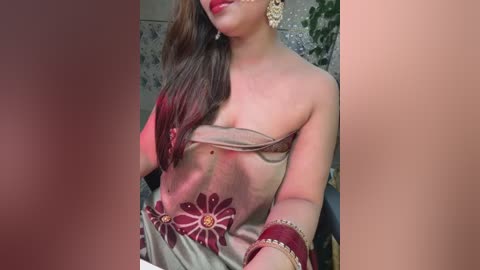 Media: Video of a South Asian woman with light brown skin, wearing a beige saree adorned with red floral embroidery. She has long brown hair, red lipstick, gold earrings, and a gold bracelet. Background features green foliage.