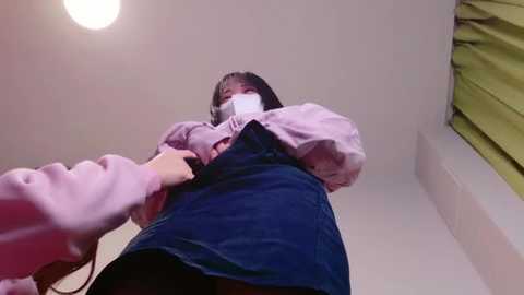 Media: Video of a young Asian woman in a pink mask, blue denim skirt, and pink sweater, taken from a low angle, indoors, with a yellow curtain in the background.