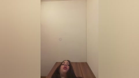 Media: Video of a woman with long dark hair and red lipstick, leaning over a wooden table in a narrow, dimly-lit room with beige walls and a small, circular light fixture on the ceiling.