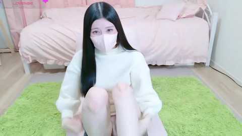 Media: A video of a young East Asian woman with long black hair, wearing a white mask and white sweater, sitting on a green rug in a pastel pink bedroom with a bed.