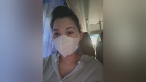 Media: Video of a woman with medium skin tone, dark hair tied back, wearing a white face mask and light grey shirt, seated indoors.