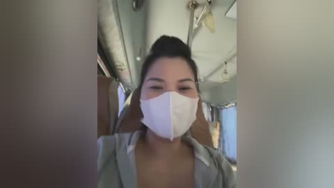 Media: Video of an Asian woman with medium skin tone, wearing a white surgical mask, light gray shirt, and black hair tied up. She is inside a dimly lit bus, with visible pipes and ventilation ducts on the ceiling.