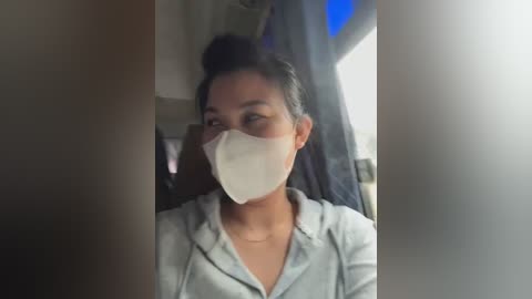 Media: Video of an Asian woman with dark hair in a bun, wearing a light grey hoodie and white face mask, sitting in an airplane cabin.