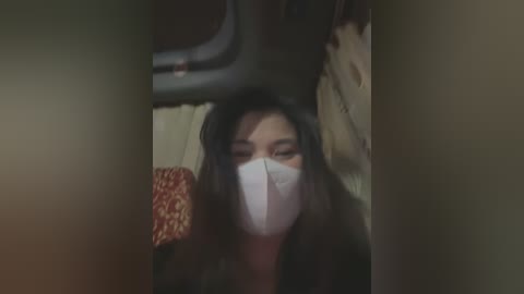 Media: A blurry video of a woman with long black hair and a white mask, sitting in a dimly lit vehicle, possibly a car or bus.