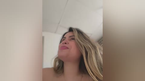 Media: Video of a smiling, light-skinned woman with long, wavy blonde hair, wearing pink lipstick, looking upward in a white room with a ceiling fan.