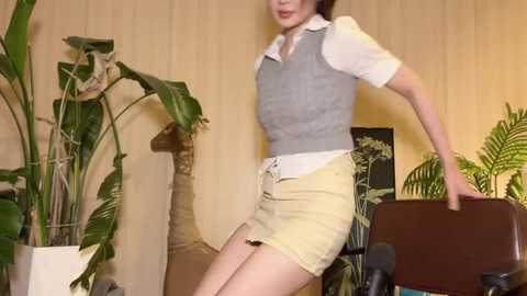 Media: Video of a slender woman in a gray vest over a white shirt, beige shorts, and black high heels, leaning on a chair, with a microphone in front. Background includes green plants and a wall with vertical wood paneling.