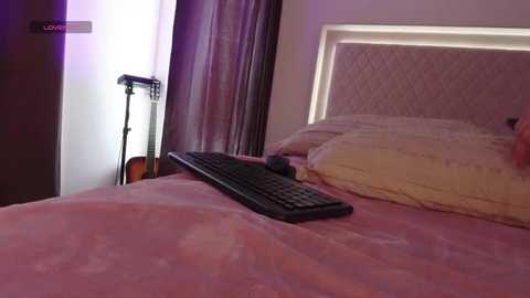 Media: Video of a cozy bedroom with a pink quilted bedspread, a beige pillow, a black keyboard, and a guitar on the floor, soft lighting, and a dimly lit \"LOVE\" sign in the background.