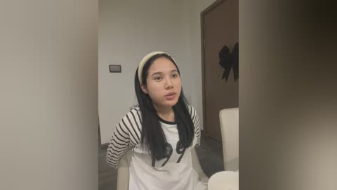 Media: Video of a young Asian woman with long black hair, wearing a white T-shirt with black stripes and a beige headband, sitting in a modern room with beige walls and a dark doorway.