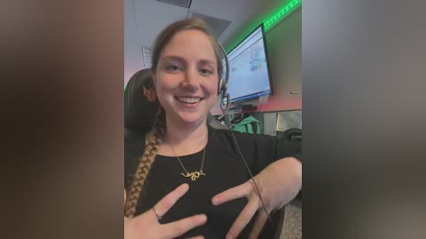 Media: Video of a smiling young woman with fair skin, brown hair, and a black top, wearing a headset, seated in a gaming chair with green LED lights, in a dimly lit room with a monitor and gaming setup.