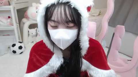 Media: A video of a young Asian woman in a white mask, red Santa hat, and red velvet cape, seated indoors with pink gaming chair, white shelves, and plush toys in the background.