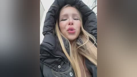 Media: A video of a young woman with long blonde hair, wearing a black puffy jacket, making a kissy face, taken from a low angle.