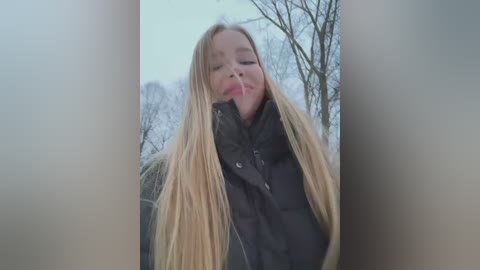 Media: Video of a blonde woman with long hair, wearing a black puffy jacket, outdoors in a wintry forest. She has her eyes closed and is puckering her lips.