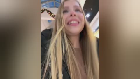 A video of a young woman with long, straight blonde hair and fair skin, smiling broadly, wearing a black jacket. The background is blurry, possibly an indoor setting.