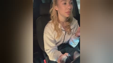 Media: Video of a young woman with wavy blonde hair, wearing a white hoodie, driving a car at night. She has a neutral expression, hands on the wheel. The background is dark, suggesting a dimly lit interior.