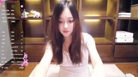 Media: Video of an East Asian woman with long black hair, fair skin, and a slim figure, wearing a white blouse, sitting at a wooden desk. Background features wooden shelves with decorative elephants and books.