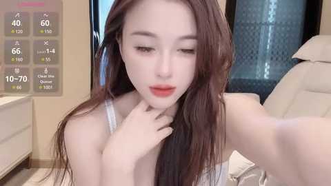 Media: Video of a young East Asian woman with fair skin and long brown hair, wearing a white tank top, lying on a bed. Background shows a modern bedroom with a white dresser, a window, and digital health stats overlay.