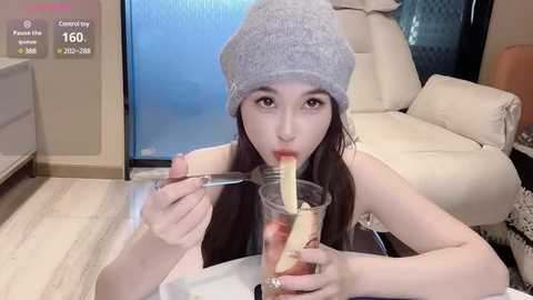 Media: Video of a young Asian woman with long hair, wearing a gray beanie, eating a strawberry from a glass, seated at a wooden table in a modern living room with cream-colored furniture.