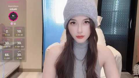 Media: A video of a young East Asian woman with long dark hair, fair skin, and a gray beanie, wearing a white top, seated indoors with a blue glass door in the background.