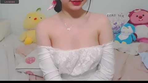 Media: Video of an East Asian woman with light skin, wearing a white off-shoulder lace dress, sitting on a bed with plush toys, including a yellow duck, pink bunny, and blue dog.