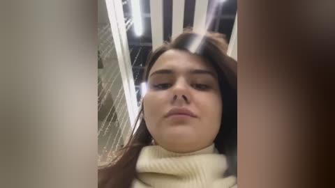 Media: Video of a young woman with long brown hair, wearing a cream sweater, looking straight ahead. Background features a subway train with vertical metal bars and fluorescent lights.