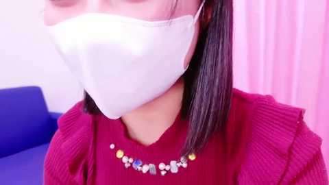 Media: Video of an Asian woman with straight black hair, wearing a white surgical mask, red sweater, and a necklace with colorful beads, seated indoors with a blue couch and pink curtains in the background.