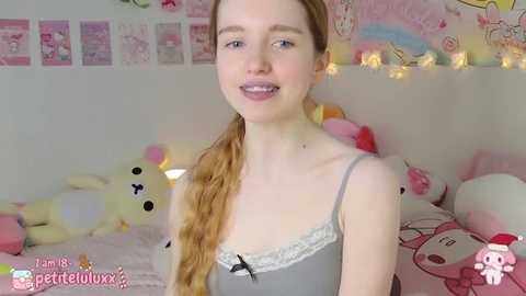 Media: Video of a slender, fair-skinned, young woman with long, light-brown hair in a braid, wearing a grey camisole, smiling in a pastel-themed bedroom with plush toys and string lights.