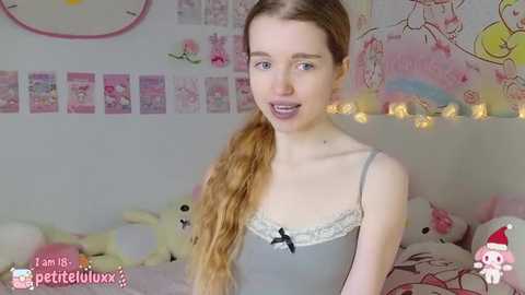 Media: Video of a young woman with fair skin, long blonde hair in a ponytail, wearing a gray camisole with lace trim, sitting in a cozy bedroom with pink decorations, including stuffed toys and fairy lights.