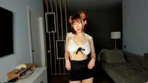 Media: Video of an Asian woman with red hair and a floral headband, wearing a white bikini top and black mini-skirt, posing in a modern living room with gray walls and a sofa.