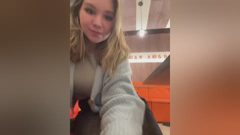 Media: Video of a young Caucasian woman with shoulder-length blonde hair, fair skin, and a light gray sweater, smiling slightly. Background features a modern, orange-themed interior with visible text in Cyrillic.