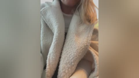 Media: A video of a woman in a cozy, cream-colored, fluffy bathrobe with her blonde hair visible. The background is blurred, suggesting an indoor setting with soft, warm lighting.