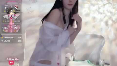 Media: Video of a young woman with fair skin, dark hair, and large breasts, wearing a white off-shoulder dress, in a softly lit, serene room with a white chair and a plant.