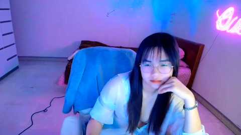 Media: Video of an Asian woman with long black hair, wearing glasses, a light shirt, and a bracelet, sitting on a bed with a blue blanket, in a room with purple and blue lighting, and a glowing butterfly wall decoration.