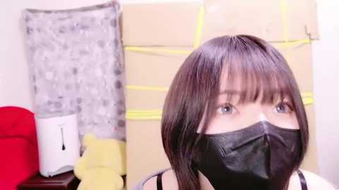 Media: Video of a young woman with shoulder-length black hair, wearing a black face mask and a white top, standing in a modern office with beige cubicles, a white printer, and a red cushion.