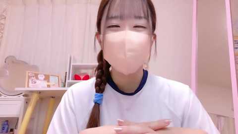 Media: A video of an Asian woman in a white mask and long brown braid, wearing a white shirt, sitting indoors with white curtains, a bookshelf, and a lamp in the background.