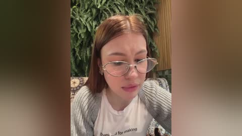 Media: Video of a young Caucasian woman with straight, shoulder-length auburn hair, wearing glasses, a grey knitted cardigan, and a white shirt. She has a neutral expression. Background features lush green foliage and a wooden structure.