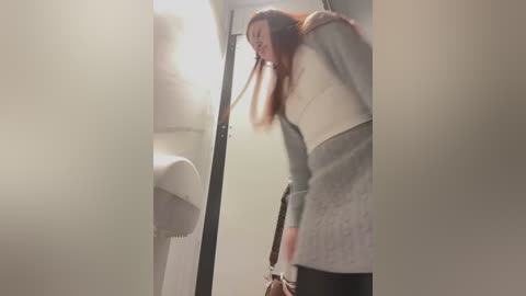 Media: A video of a woman with long red hair, wearing a gray dress and white top, leaning into a public restroom stall. The background is foggy and dimly lit, with a white paper towel dispenser on the left.